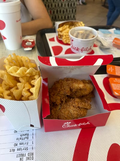 This image displays an indoor scene within a Chick-fil-A restaurant, located in El Paso, United States. The foreground features a meal set on a light-colored table. An open red and white Chick-fil-A box contains multiple breaded chicken strips, positioned next to a white patterned carton overflowing with waffle fries. A partially visible receipt underneath the fry carton lists 'Strips 4ct,' 'Fries MD,' 'Buffalo,' and 'Ranch.' A white Chick-fil-A beverage cup with a lid and a small red Heinz ketchup bottle are also on the table.
On a black tray in the mid-ground, another portion of waffle fries, a lidded white Chick-fil-A cup containing an unclear dark item, and multiple small orange plastic dipping sauce containers are arranged. An individual's arm and patterned torso are visible on the left, indicating a diner. In the blurred background, a person's lower legs in blue pants are partially discernible. The environment is a typical brightly lit fast-food dining area.