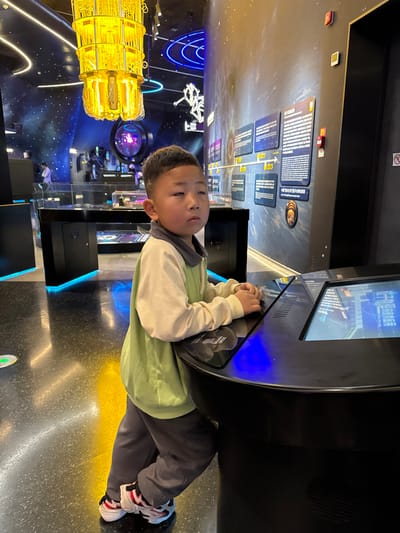 The image depicts the interior of a modern exhibition hall in Guangming District, China. A young East Asian boy, approximately 5-7 years old, stands in the foreground, angled slightly to his right and facing forward. He wears a long-sleeved top with a light green torso and cream-colored sleeves, dark grey pants, and white athletic shoes with black and red accents. The boy is leaning against a dark, curved interactive display kiosk, with his hands resting on its glossy black surface. The kiosk features at least one illuminated screen displaying blue graphics and Chinese text. He maintains a direct gaze with a neutral expression.

The background reveals a high-tech exhibition environment. Dark walls are adorned with a space-themed mural depicting stars and galaxies, alongside multiple wall-mounted information panels displaying Chinese text and diagrams in a timeline format. Numerous dark display tables with clear tops showcasing exhibits and featuring blue underlighting are visible throughout the space. A prominent, multi-tiered, yellow-orange chandelier, constructed from intricate components, hangs from the ceiling. Blue LED strip lighting accentuates architectural elements and outlines structures. The floor is dark, reflective, and speckled. Other individuals are present in the distant background, out of focus.
