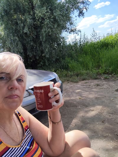 A woman with blonde hair holds a red mug with a white rim, displaying text. She is wearing a brightly colored striped tank top and a necklace. A bracelet adorns her arm. In the background, a silver car is partially visible, along with lush green foliage and trees under a partly cloudy sky. The ground is a mix of dirt and dry leaves. The woman's expression appears serious. The lighting suggests it is daytime. The text on the mug reads "GRANDDAD THE KING OF THE WORLD".