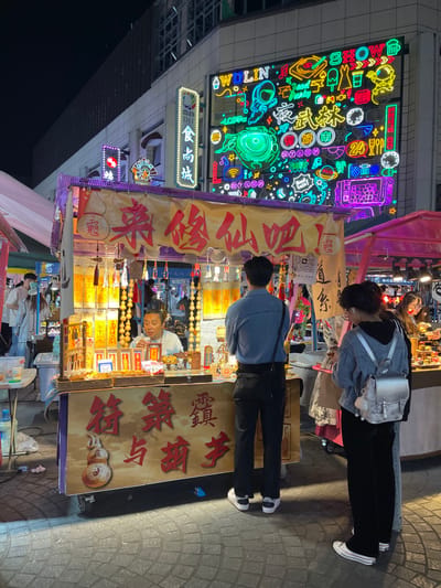 The scene is a street stall in Hang Zhou Shi, China, located at 30.264 latitude and 120.156 longitude. The stall is brightly lit and decorated with colorful lanterns and signs with Chinese writing. The stall is selling various trinkets and souvenirs, including small, brightly colored figures. There are three people in the scene: a man behind the counter, a man in front of the counter, and a woman standing to the right. The man behind the counter is sitting and appears to be selling something to the man in front of the counter. The woman is standing with her back to the camera, observing the scene. The environment is crowded, with other stalls and people in the background. The buildings in the background are illuminated with brightly colored neon signs. The scene is dynamic and bustling with activity.