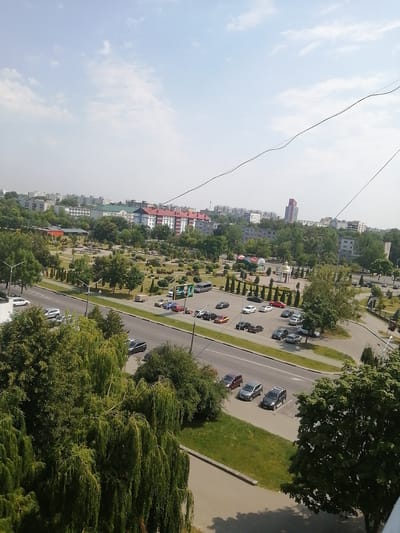 The image shows an aerial view of a park and residential area in Salihorsk, Belarus. A wide road with several cars is visible in the foreground, with a park and green spaces beyond. Buildings and trees are scattered throughout the scene, and a tall, modern building stands out in the distance. The sky is partly cloudy, and the overall atmosphere suggests a typical day in a suburban environment.