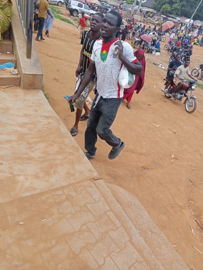 A man in a white and red jersey with a star emblem walks down a dirt path in a bustling outdoor market in Sauka, Nigeria. He carries a white bag and a green bottle in his hand. Behind him, another man in a black and yellow striped shirt walks with a slight limp. In the background, a crowded market scene unfolds with numerous motorcycles, stalls, and people milling about under umbrellas. The ground is a mixture of dirt and some paved areas.