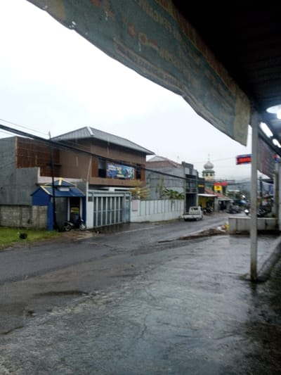 The image displays an urban street scene in Chagzam_Pam, Bhutan, characterized by an overcast sky and wet asphalt surfaces, indicating recent rain. A large, dark banner with indecipherable text hangs from an overhead structure, partially covering the upper left quadrant.
The street, appearing wet and reflective with puddles, extends from the lower left towards the mid-right. On the left side of the street, a series of structures are visible: a low concrete wall, followed by a small, standalone blue kiosk with a yellow roof trim, in front of which a dark-colored scooter is parked. Adjacent is a two-story building with a brown facade and a prominent peaked roof, featuring a blue and yellow advertisement banner on its upper level. Further along is a building with a light grey, vertically slatted gate, and beyond it, additional residential or commercial structures.
On the right side of the street, a white utility pole stands in the midground, supporting electrical lines and a light fixture. Below an unseen overhang, a series of illuminated fluorescent lights are visible. A white pickup truck is positioned on the wet road in the mid-distance, facing away from the camera. Further down the street, buildings line both sides, culminating in a distinct building with a green-domed minaret visible in the background, slightly right of center. Hazy mountain outlines are discernible on the horizon beneath the uniformly grey sky. No individuals are visible within the frame.