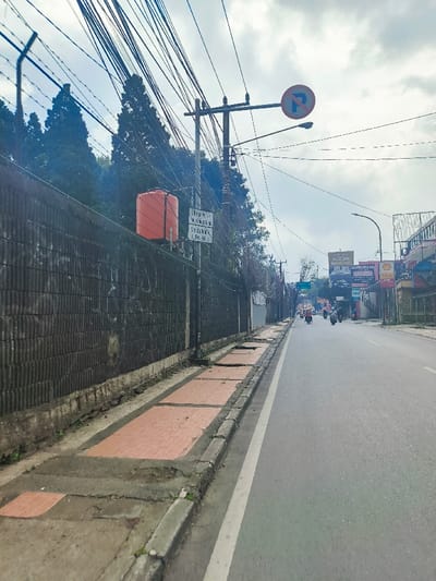 The scene is a street in Lembang, Indonesia, located at latitude -6.816 and longitude 107.616. It is a paved, two-lane road with a sidewalk on the left side, viewed from a position on the sidewalk, facing the opposite direction of traffic. The road is relatively empty with a few vehicles visible, including a motorbike, and a few figures in the distance. The sidewalk has a red tile pattern, but there are some broken sections and missing tiles. On the left side of the street, there is a high, black, vertical wall made of corrugated metal. There are multiple power lines stretching across the image, along with several utility poles. A bright orange-red water tank is mounted on a pole, located in the center of the image, beside a sign with text in Indonesian. A street sign with a white "P" in a blue circle on a white background is visible on a pole across the street. The sky is overcast, with some small clouds visible.