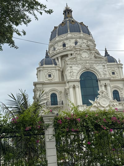 The image displays the exterior of a large, ornate building with multiple domes, reminiscent of classical architecture. The building is predominantly white with dark blue domes topped with gold accents. It features intricate carvings, columns, and arched windows. In the foreground, a black wrought-iron fence is covered with lush green foliage and small pink flowers, obscuring the lower part of the building. Palm fronds are visible to the left, and a large tree with dark green leaves hangs from the upper left corner. The sky is overcast. The building is located in Hoa Lư Ward, Vietnam. There are no people visible in the image.
