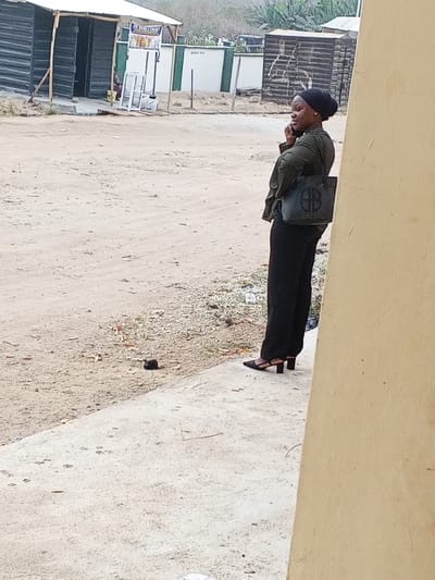 A young woman stands on a concrete sidewalk, talking on a cell phone. She wears black pants, a dark green shirt, black heels, and a head covering. A black purse hangs from her shoulder. The background features a dirt and sand-covered area, buildings with dark walls, and some greenery in the distance. The composition is somewhat obstructed by a wall in the right foreground.