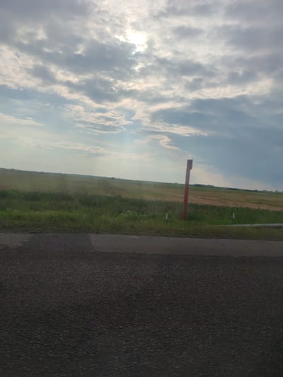 The image depicts an outdoor scene with a cloudy sky and a vast agricultural field. In the foreground, a dark asphalt road is visible. Beyond the road, a strip of green grass leads to a large, light-brown field that appears to have been harvested. A tall, red-and-brown post stands in the grassy area, likely marking a boundary or utility. The sky is filled with dramatic, diffused sunlight breaking through heavy, grey clouds, creating streaks of light. The environment suggests a rural setting in Аздзелінскі сельскі Савет, Belarus, possibly during late summer or early autumn.