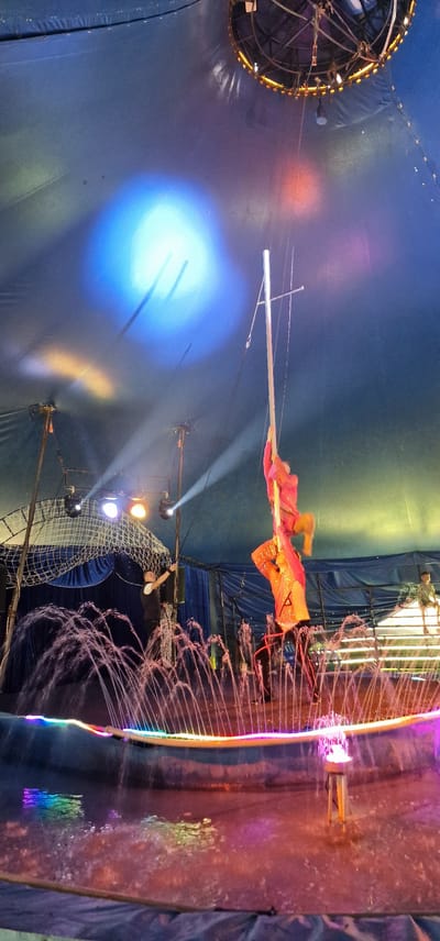 The image depicts a circus tent interior with a performance underway. The ceiling of the tent is a dark blue fabric, illuminated by spotlights creating large circular and oval patterns of blue and yellow light. A circular light fixture with yellow bulbs is visible at the apex of the tent.
In the center of the frame, two performers are engaged in an aerial act. One performer, wearing a bright orange and sequined costume, is positioned higher up on a vertical pole, with their legs wrapped around it. Another performer, also in a similar bright costume, is below, holding onto the pole and the upper performer. Their poses suggest they are in motion.
Below the aerialists, a circular fountain is active, with numerous arcs of water spraying upwards. The rim of the fountain is illuminated by colorful LED lights, cycling through pink, blue, and yellow. The water in the fountain reflects these lights.
On the ground level, to the left, a man in a dark vest and white shirt stands near some stage equipment, possibly a lighting rig, and points upwards. To the far right, a child in a yellow shirt is visible sitting on a raised platform. The overall environment suggests a circus performance setting with a water feature as part of the show. There is no specific indication of the city of Saulkrasti, Latvia, within the image itself.