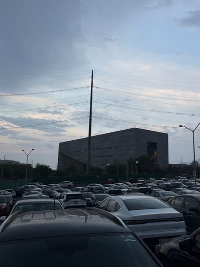 The image depicts an expansive outdoor parking lot in Ciudad Santa Catarina, Mexico, at dusk under a heavily clouded sky transitioning from pale blue to dark grey. The foreground is dominated by rows of densely packed vehicles, primarily sedans and SUVs of various colors. A dark-colored vehicle occupies the immediate foreground, showing only its roof and windshield. Directly behind it, a white BYD sedan is parked facing away from the viewer, its rear visible with a partial license plate "PRF-62-".
In the midground, a dark green security fence separates the parking area from the background. Beyond this fence stands a prominent, tall, slender power transmission tower, dark grey, with multiple power lines extending horizontally across the frame. To the right and behind the tower, a large, multi-story, modern building constructed of dark grey concrete dominates the skyline. This brutalist-style structure features horizontal window bands and a significant rectangular recessed opening on its visible facade. One side of the building exhibits a sloped, textured surface. Several streetlights with dual luminaires are positioned throughout the parking lot, with one on the far right displaying a vertical "F" sign. No individuals are visible.