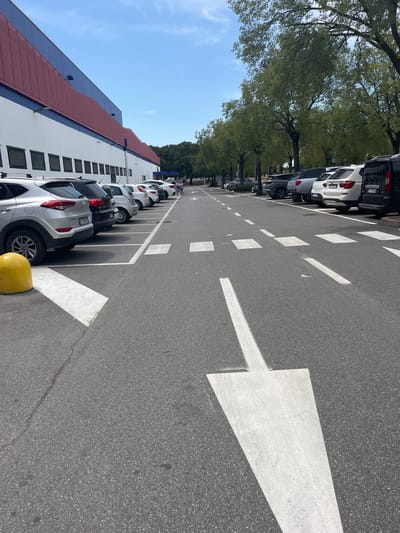 The image shows a parking lot in front of a large building with a red and blue facade. The building appears to be a commercial establishment, possibly a supermarket or warehouse. The parking lot is asphalt, with white lines marking parking spaces and directional arrows. Several cars are parked in the spaces, and some trees line the edge of the parking lot. In the distance, two people are pushing a shopping cart. The sky is blue with some clouds. This scene is consistent with the parking area of a large retail store, such as a Costco, located in Castiglione delle Stiviere, Italy.