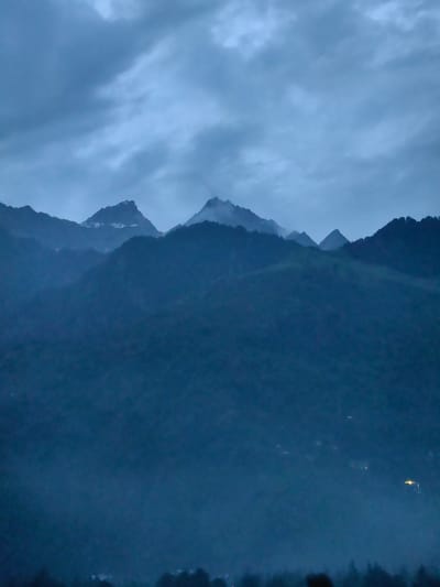The image depicts a mountainous landscape under a cloudy, twilight sky. The foreground is dominated by dark, densely forested mountain slopes, partially obscured by mist. In the mid-ground, layers of receding mountains are visible, with their shapes softened by the atmospheric haze. The peaks of the further mountains are sharper, with some patches of snow or lighter rock visible on their summits. The sky is filled with heavy, dark grey clouds, suggesting an overcast or approaching storm. Scattered lights, appearing as small yellow or white dots, are visible in the lower sections of the mid-ground and foreground mountains, indicating human settlements or structures. The scene is devoid of people or discernible actions. The overall impression is one of a vast, natural, and somewhat somber environment, characteristic of a mountain region. The provided location of Bahang, India, would place this scene within the Himalayan mountain range.