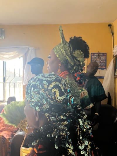 In an indoor setting in Warri, Nigeria, three individuals are present. The woman on the left wears a green head wrap, while the woman on the right has a black head covering decorated with pale green sequins. The man in the background is partially visible, wearing a dark green shirt with a colorful patterned design. A window is visible in the background, letting in bright sunlight. The wall is painted a muted yellow. A framed object hangs on the wall behind the man.
