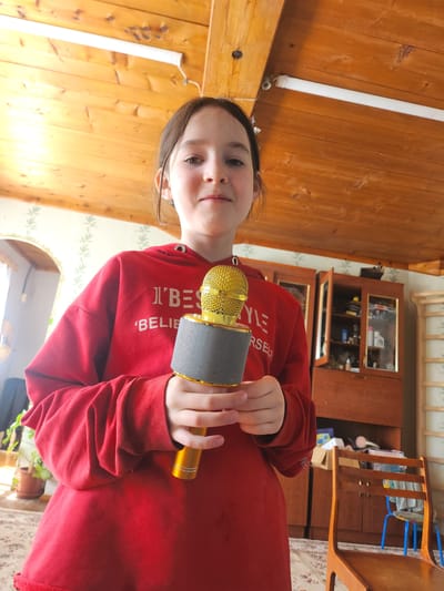 A young girl stands in a room with wood-paneled walls and ceiling, holding a gold and gray microphone. She is wearing a red hooded sweatshirt with white text that reads "I BELIEVE IN YOURSELF". Behind her is a wooden cabinet with glass doors. The room contains a wooden chair with blue accents. The location is Архангельское, Russia.