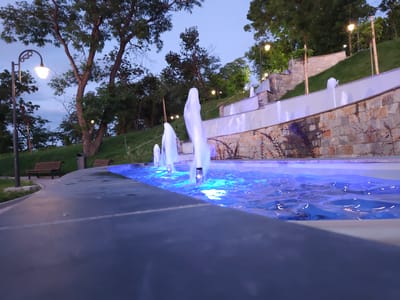 The image depicts a beautifully lit tiered fountain in the city of Svishtov, Bulgaria, likely in a park or public space. The fountain is illuminated with vibrant blue lights, creating a striking visual contrast against the twilight sky and the surrounding greenery. Water jets arch gracefully from the different levels of the fountain, adding dynamism to the scene. Stone walls and pathways form the structure of the fountain, with lush trees and grass visible in the background. Several lampposts with vintage-style lamps are also scattered throughout the area, enhancing the ambiance. Two park benches are visible, suggesting a place for public relaxation and enjoyment. The low-angle perspective emphasizes the grandeur of the fountain and the interplay of light and water.