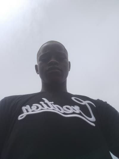 A low-angle shot depicts a young Black male wearing a black t-shirt with white lettering. The sky is overcast. There is no clear evidence that this photo was taken in Epe, Nigeria.