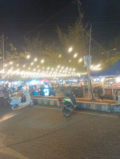 This is a night-time photograph taken at the Luwoo beach street food area in Luwoo, Indonesia. The scene depicts a bustling outdoor dining environment with numerous stalls illuminated by strings of fairy lights suspended above. Several people are visible, seated at tables and congregating around food stalls. In the foreground, two scooters are parked on the asphalt road. One scooter is light blue, and the other is dark green. The background features tents and trees. A sign with text is mounted on a pole in the middle ground. The overall atmosphere is lively and social.