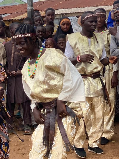 The image captures a group of people, predominantly young men and boys, gathered outdoors in what appears to be a rural setting in Rigachikun, Nigeria. The scene is bustling, with many individuals visible, some in the foreground and others in the background.
Several individuals are dressed in traditional attire. Some wear elaborate, light-colored robes with intricate patterns, possibly for a ceremony or special occasion. One person in the foreground is wearing a dark brown garment with embroidered details and a headband adorned with shells. Another person has a colorful beaded necklace. Some individuals are wearing black shoes, while others are barefoot or wearing sandals.
Objects of interest include what appear to be leather whips or sashes with fringe, held by some of the men. The background shows hints of a rustic environment, with corrugated metal roofing visible, suggesting a simple, possibly impoverished, community. The ground is dusty earth.
The overall mood seems to be one of communal gathering, possibly a celebration or a religious event. The expressions on the faces of the people range from focused to smiling. The lighting suggests it is daytime, with bright sunlight casting shadows. No visible text is present in the image.