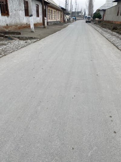 The scene depicts a straight section of a newly paved, light gray asphalt or concrete road in Akhtachi, Uzbekistan, extending into the distance under an overcast sky. The road surface exhibits minor dark specks and a few subtle linear marks.
On the left side of the road, an unpaved shoulder of dark earth and loose gravel separates the roadway from a row of single-story buildings. The nearest building features a textured, off-white facade, a reddish-brown base trim, and three visible rectangular windows with brown frames. Beyond it, another single-story structure with a light beige or yellow facade and reddish-brown window trim is visible. Utility poles with overhead wires run along this side of the street.
On the right side of the road, a shoulder appears to be under construction, consisting of a fresh layer of light gray crushed stone or gravel adjacent to the pavement, followed by darker exposed earth. Further down the road, several single-story buildings with light-colored walls and dark, sloped roofs are visible, interspersed with numerous tall, slender, bare deciduous trees.
In the far distance, a white van, possibly a minivan, is parked or traveling slowly on the right side of the road, facing away from the observer. Two individuals are discernible on the left side of the road in the distance. One individual, dressed in dark clothing, is near a blue object. The overall environment suggests a residential or semi-rural setting during a non-foliage season.