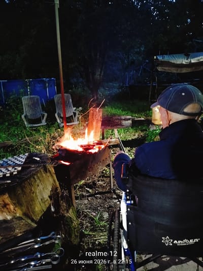 An elderly man in a wheelchair, wearing a blue jacket and a baseball cap, sits facing a bonfire in a backyard at dusk. The fire is burning in a metal brazier on the ground, with several skewers of meat placed above it on a wooden rack. Two empty plastic chairs are visible behind the brazier. A large tree with dark foliage stands in the background, and beyond that, a blue tarp is stretched over a structure. The ground is covered in grass and leaves. The image has a timestamp overlay indicating "realme 12" and "26 июня 2026 г. в 22:15". The location is identified as Novoe, Russia.