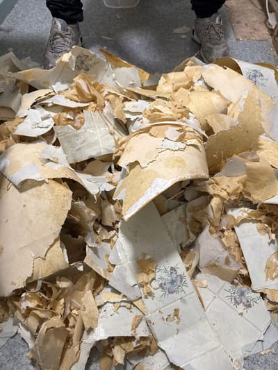 The image shows a pile of torn-off wallpaper on the floor. The wallpaper appears to be old and has a floral pattern. Two feet wearing athletic shoes are visible at the top of the image, suggesting someone is standing over the pile of debris. The floor is a speckled gray color, possibly linoleum or concrete. This scene could be found in a residential or commercial property undergoing renovation within the town of Hemel Hempstead, which is located in the borough of Dacorum, United Kingdom.