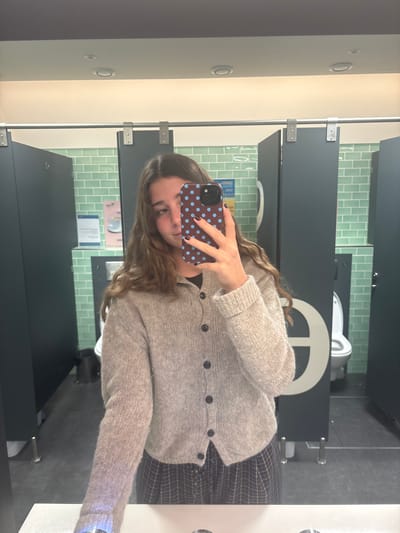 The image depicts a mirror selfie taken by a young individual inside a public restroom located in Pombal, Portugal.
The central subject is a person with long, wavy brown hair, wearing a light brown knitted cardigan with four visible dark buttons over a dark top, and a dark plaid skirt. Their right hand holds a smartphone with a dark blue case adorned with light blue polka dots, positioned to capture their reflection. Dark nail polish is visible on the fingers of the right hand, which partially obscures the subject's face.
The restroom environment features light green rectangular tiles arranged in a brick pattern on the walls. Several dark grey or black toilet stalls are visible, equipped with silver-toned metal hinges and latches. Inside the rightmost visible stall, a white toilet with its seat in the upright position is discernible. A white countertop, presumably part of a sink vanity, occupies the bottom foreground. The ceiling is light-colored and fitted with two visible recessed lighting fixtures. The floor is dark grey. A white circular graphic is partially visible on the door of the rightmost stall, and a faint poster is present on the tiled wall between two stalls.