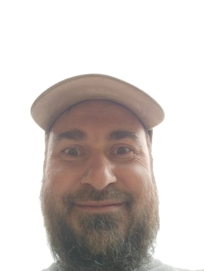 A man wearing a light-colored baseball cap is pictured from the chest up, looking directly at the camera with a slightly surprised expression. He has a full beard and is wearing a grey t-shirt. The background is a bright, blown-out white, suggesting an outdoor setting or bright lighting conditions. The image appears to be a selfie taken outdoors, possibly in the village of Shiryayevo in the Ilyinsky district of Russia, given the context provided.