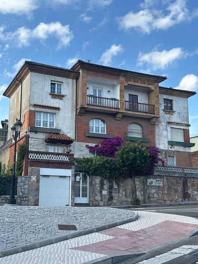 This media file presents a rotated view of a building and an adjacent street in Oviedo, Spain, under a bright blue sky with scattered white clouds. The image is oriented such that the sky occupies the left side, the building extends horizontally across the middle, and the street and sidewalk are positioned on the bottom right.
The multi-story building is characterized by its layered facade, incorporating weathered white stucco, sections of red brick, and rough-hewn grey stone. It features numerous windows with both dark and white frames, some equipped with closed white shutters or blinds. Two wooden balconies with decorative railings protrude from the building's upper sections. A striking element is the vibrant purple bougainvillea plant, intertwined with green foliage, appearing to hang downwards from the building's exterior (indicating its vertical growth in reality). Other notable architectural details include a small arched window set in a brick section, a small tiled awning over another window, and a white air conditioning unit attached to the lower stone section.
The right side of the image reveals a distinctive street or sidewalk area. It is paved with grey and reddish-pink tiles arranged in a prominent diagonal diamond or arrow pattern. This patterned surface borders a grey curb, which in turn runs alongside a stone wall. Affixed to this stone wall are two small square plaques; one displays the number "2" and other faint symbols, while the other features a stylized "S" or "5" within a circle. Further along the wall, a partially obscured sign with a red circular outline and a diagonal line can be seen. A black street lamp with an ornate design is visible in the bottom-left corner of the frame.
The bright lighting and clear sky indicate a sunny daytime setting. No individuals are present in the scene.