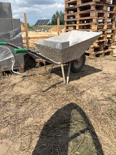 The image depicts an outdoor construction site in Opatów, Poland. A wheelbarrow, filled with a single concrete block, is positioned in the foreground. To the left of the wheelbarrow, stacks of grey concrete blocks are visible, wrapped in clear plastic sheeting. In the background, a large stack of wooden pallets stands to the right. Beyond the pallets, solar panels are installed on a sloped surface. The ground is covered with dry grass and loose soil. The scene is illuminated by daylight, casting a shadow of the wheelbarrow onto the ground.