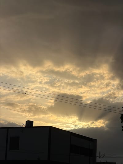 The image depicts a dramatic sky with crepuscular rays beaming through dense clouds, illuminating a portion of the horizon. A silhouetted building with a flat roof and visible air conditioning unit dominates the lower portion of the frame. Overhead, power lines are strung across the scene, with two small birds perched on one of them. The overall lighting suggests either dawn or dusk, creating a somber and atmospheric mood. The location is identified as Maracay, Venezuela.