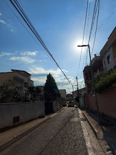 The image shows a narrow, paved street in Tiranë, Albania. The street is lined with buildings on both sides, with a yellow car parked on the right side of the road. There is a streetlight on the right side of the street with numerous power lines stretching across the sky. The sun is shining brightly, casting shadows on the street. The sky is blue with a few white clouds. There are bushes and trees lining the edges of the street. The image appears to have been taken from the middle of the road, facing towards the sun. The image was taken at the location of 41.336, 19.803 in Tiranë, Albania.