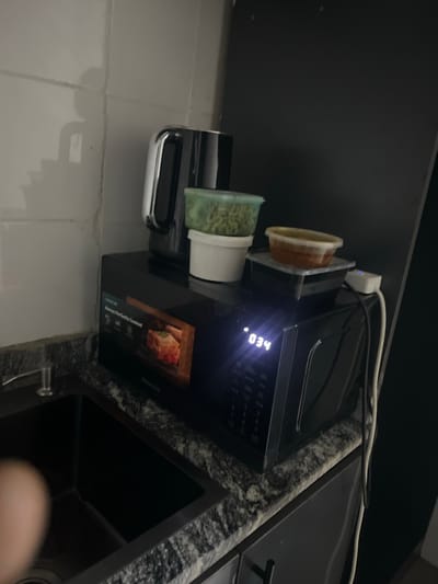 A black Hisense microwave oven sits on a granite countertop next to a black sink. The microwave displays "034" and an image of food. On top of the microwave are two containers of food, one green and one orange. A kettle is to the left of the food containers. The background is a white tiled wall with a shadow on it. The scene is in a kitchen in Karum, Nigeria.