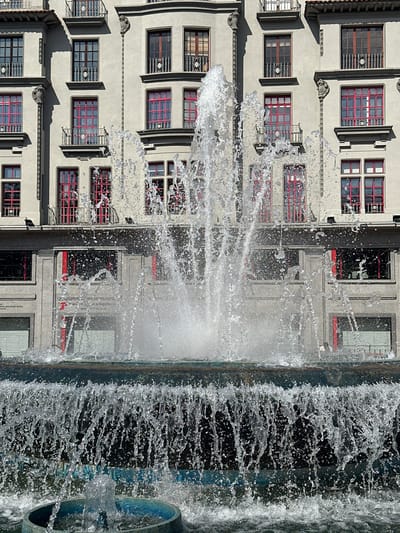 The image shows a close-up, vertical view of a building facade adjacent to a cascading fountain. The building appears to be several stories high, with repeating architectural elements including numerous windows. The windows are rectangular with red frames and a grid pattern on the interior. Some windows have decorative metal grates. The facade is a light-colored, likely stucco, material.

The fountain occupies the right side of the frame and is in motion, with water spraying and falling. The water creates a dynamic, blurred effect, contrasting with the sharp lines of the building. The fountain appears to be a constructed water feature, possibly part of a public space or architectural design.

The time of day appears to be daytime, as evidenced by the bright lighting and shadows cast on the building. The weather seems clear and sunny. There are no people visible in the image. The architectural style suggests an urban setting, possibly in a European city.

One of the windows, partially visible on the lower right of the building in the original image, has text that appears to read "INSTITUTO NACIONAL DE PREVISIÓN" and below that "OFICINA DE", followed by more text which is obscured. This suggests the building might be a public institution. The context of Oviedo, Spain, is provided.