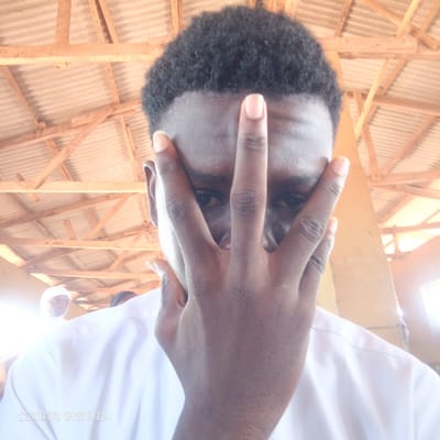 The image depicts a close-up of a young man in Edu Town, Nigeria, partially obscuring his face with his hand. He has dark, curly hair and is wearing a white collared shirt. His right hand is positioned in front of his face, with his fingers spread and touching his forehead and cheeks, revealing only his eyes and forehead. The background shows a wooden structure with corrugated metal roofing, suggesting an outdoor or semi-covered area. Sunlight is filtering through, creating bright spots and lens flares, indicating it is daytime. In the background, another person wearing a white head covering is visible, along with parts of other individuals. The overall lighting is bright. There is text visible at the bottom left corner of the image: "IXDEN DIGITALS".