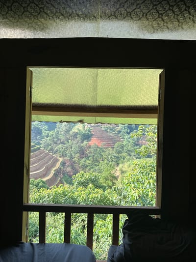 A view from a dark interior space through a framed window shows rice terraces, foliage, and soil in the Hoàng Su Phì Commune, Vietnam. The interior features patterned glass on the left, a wooden window frame, and bedclothes on the right.