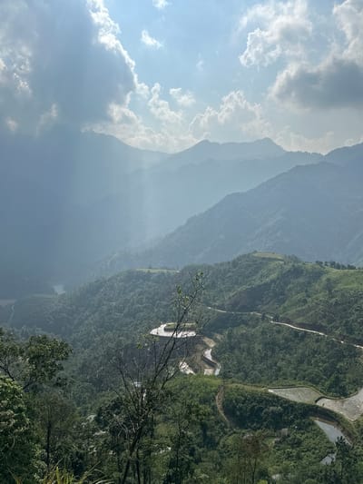 This image presents a stunning, expansive mountainous landscape within Thông Nguyên Commune, Vietnam, captured from a high vantage point looking into a vast valley. The scene is characterized by layers of mountains, lush vegetation, and agricultural terraces under a bright sky.

In the upper portion, a clear blue sky is adorned with scattered white and grey clouds, through which soft sunlight streams from the left, illuminating the scene. Below the sky, successive mountain ranges recede into the distance, each layer appearing softer and more blue-grey due to atmospheric haze, creating a profound sense of depth.

The closest mountain slopes, particularly on the right side of the frame, are densely covered with vibrant green foliage, indicative of rich forests. A light-colored, winding path or road can be seen traversing one of these verdant slopes. Within the middle ground, distinct terraced fields, likely rice paddies, are visible. Some of these terraces are filled with water, reflecting the sky and exhibiting organic, curving shapes that conform to the landscape. Scattered among the trees and near the terraced areas are small structures, including a noticeable white building complex.

The immediate foreground, particularly towards the bottom right, features dark, silhouetted tree branches and leaves, partially framing the panoramic view and emphasizing the scale of the distant landscape.

The time of day appears to be daytime, possibly morning or afternoon, given the bright illumination. The weather is clear with some cloud cover, and an omnipresent atmospheric haze softens the distant features. No people are visible, and there is no discernible text in the image. The overall impression is one of serene natural beauty, showcasing the grandeur of the mountains and the intricate patterns of human agricultural activity in harmony with the environment.