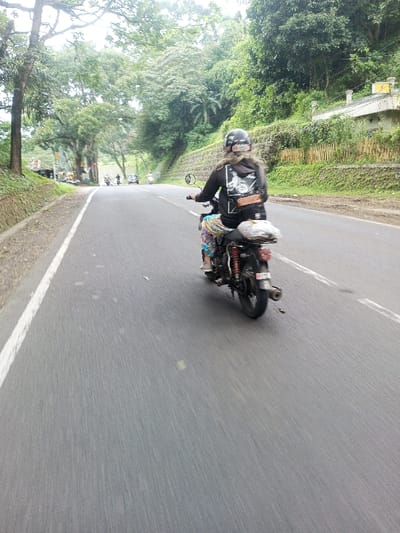 An individual operates a dark-colored motorcycle on a paved two-lane road in Khajuripada, India. The motorcycle is equipped with red rear shock absorbers and a rear exhaust pipe on its right side. A white object or bag rests on the rear seat. The rider wears a black helmet with white markings, a black long-sleeved garment featuring a white graphic print on the back, and multi-colored patterned pants. Long, light-colored hair extends from beneath the helmet. A brown, leather-like pouch is secured at the rider's lower back.
The road features a solid white line delineating its right edge from the perspective of the camera. Both sides of the road are lined with dense, green vegetation and concrete retaining walls. On the right, a steep, heavily wooded hillside rises, where a white building with an orange roof and yellow signage is partially visible. On the left, trees and vegetation also line a more gradual incline. Several additional vehicles, including motorcycles and potentially a car, are visible in the distance on the road, traveling in both directions. The sky is overcast.