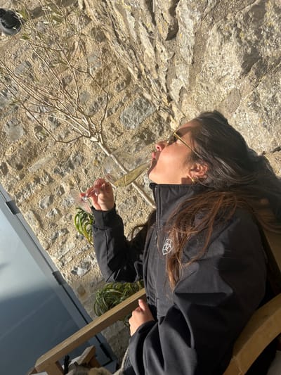 A woman with long, dark hair and glasses is sitting outside, possibly on a patio or deck, in Bristol, United Kingdom. She is wearing a black jacket. She is holding a wine glass, which appears to be filled with white wine. In the background, there is a stone wall, some bare branches, and foliage from plants. Part of a chair and what appears to be a grey, reflective surface are also visible. The scene seems to be lit by natural sunlight.