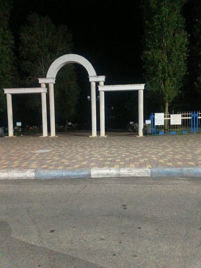 The image shows an outdoor scene at night, possibly in a park or public space. In the foreground is a paved area with a patterned surface, bordered by a concrete curb. Beyond the curb is a dark asphalt road.
Dominating the mid-ground is a white architectural structure consisting of multiple columns and archways. There are three main segments: a large central arched gateway and two flanking rectangular archways. The structure appears to be made of stone or a similar material. Behind the structure, trees are visible, their foliage dark against the night sky.
To the right of the central archway, there is a blue and white metal fence. Mounted on posts are several signs with text in a language that appears to be Cyrillic. Some of the signs have a light blue header. Near the fence, there are bushes or low-lying plants. A lamppost is visible behind one of the columns.
The overall lighting suggests artificial illumination is present, casting shadows and highlighting the architectural features and paved areas. The background is predominantly dark, indicating it is nighttime and the area may be sparsely populated. There are no people visible in the image. The location is identified as Pivdenne, Ukraine.