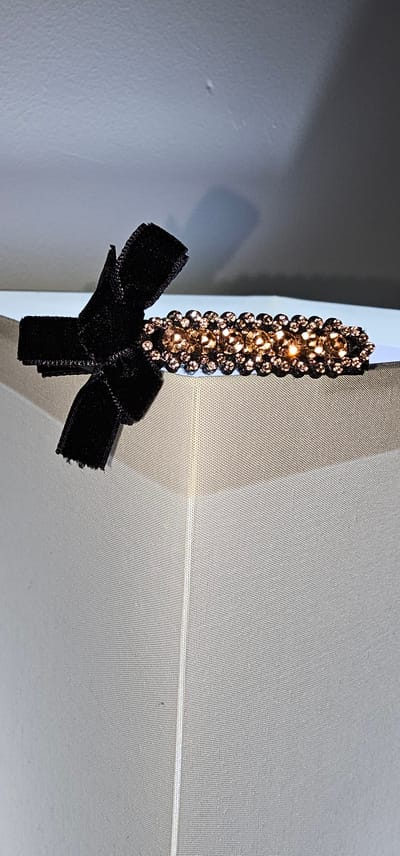 A close-up shot focuses on a decorative item, possibly a hairpin or clip, attached to a light-colored textured surface, which appears to be fabric. The item itself is designed with a vertical row of sparkling rhinestones in a warm, amber hue, arranged in multiple lines. Below the rhinestones, a black velvet ribbon is tied into a neat bow, with the ends of the ribbon trailing downwards. The contrast between the shimmering stones and the matte black velvet creates a visually appealing aesthetic.

The background is out of focus, but it suggests an indoor setting with soft lighting. The light-colored surface has a subtle weave pattern, indicating it could be a curtain, tablecloth, or upholstery. The overall impression is one of elegance and delicate craftsmanship. There are no people visible in the frame, and no specific activity is taking place. The focus is solely on the detailed ornamentation of the accessory. The lighting suggests it could be daytime, but no definitive time of day or weather cues are apparent. No text is visible in the image. The location context of Makati, Philippines, does not provide any direct visual clues within the frame itself, as the image is a close-up of a small object.