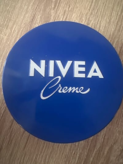 The image is a close-up, overhead shot of a round, blue Nivea Creme tin placed on a light brown wooden surface. The Nivea logo, in white, is prominently displayed on the tin. The top line of the logo reads "NIVEA" in a sans-serif font, with each letter appearing bold and raised. Below this, "Creme" is written in a flowing, script-like font, also in white and slightly embossed. The wooden surface shows a grain pattern and appears to be a table or floor. The lighting suggests an indoor environment, possibly with natural light from the side casting subtle shadows. There are no people or animals present in the image, and no specific geographical markers place this scene within Vitsebsk, Belarus.