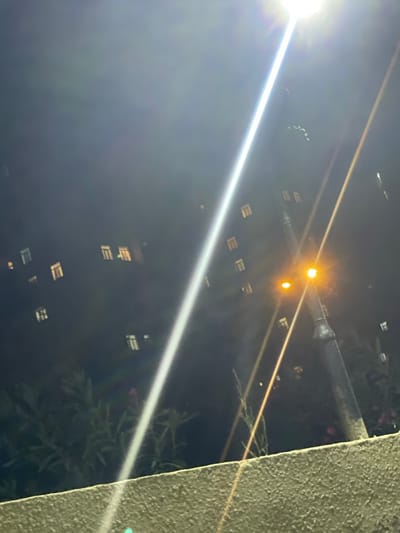 This is a nighttime photograph taken in the city of Sumgayit, Azerbaijan, depicting a dimly lit street with a prominent lamppost in the foreground. The lamppost has two lights, one brighter orange-yellow and a smaller one below it. Streaks of light from the lamppost cut diagonally across the image. In the background, a dark building with numerous lit windows is visible. Several trees and foliage are present in the lower portion of the image, with a textured, light-colored wall or ledge in the immediate foreground. The overall atmosphere is dark, with artificial lighting creating lens flare and light scattering effects.