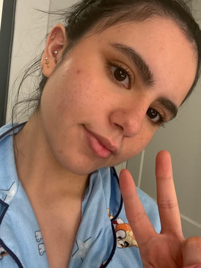 The image is a close-up, self-taken photograph of a young woman's face and upper chest. She is wearing light blue pajamas with cartoon characters and stars. Her hair is dark and pulled back loosely, with some strands falling forward. She has dark brown eyes, dark eyebrows, and visible eyelashes. There are several ear piercings on her left ear, including a small pearl stud, a hoop with a charm, and other stud earrings. Her skin shows some texture and minor blemishes. Her lips are slightly parted. She is holding up her right hand in a peace sign gesture. The background is a plain, light-colored wall. The image is taken in what appears to be an indoor setting, possibly a bedroom. No specific location in Austin, United States of America, is identifiable from the image.