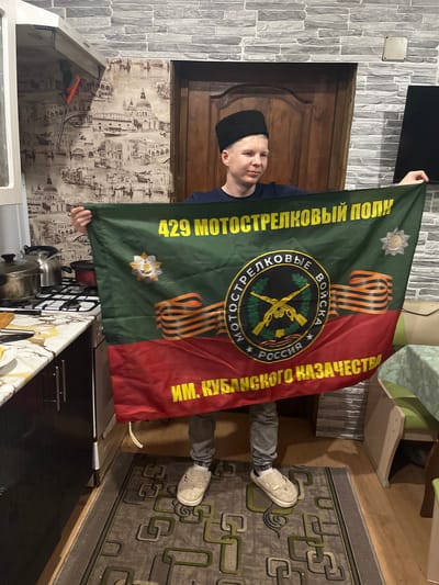 A person is holding a flag indoors. The flag is green and red with a central emblem and text in Russian, indicating it is the flag of the 429th Motorized Rifle Regiment named after the Kuban Cossack Army. The person is wearing a black hat and grey pants. The setting appears to be a kitchen. There is a gas stove, wallpaper featuring city scenes, and a patterned rug on the floor. There is a door visible behind the person and the wall appears to be made of brick. It is not possible to ascertain from the image that this is Vesyoloye, Russia.