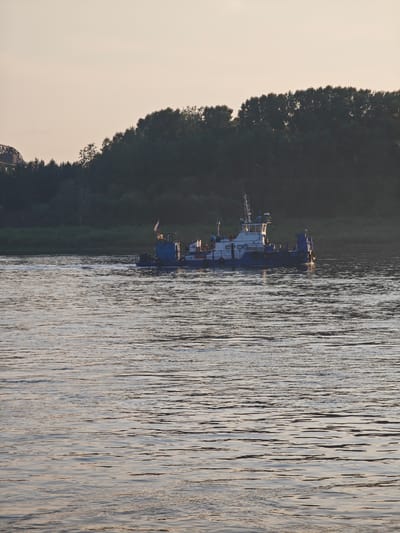 A blue and white ferry boat with a Russian flag sails on a wide, calm river at dusk. The boat has a cabin structure with windows and railings. The river is surrounded by a dense forest on the far bank, and a faint outline of a bridge can be seen on the left side of the image, suggesting an urban or semi-urban setting within or near Lesosibirsk, Russia. The sky is a pale, overcast hue, indicative of twilight. The water's surface reflects the soft light, creating ripples and streaks of shadow. There are no visible people on the deck of the boat.