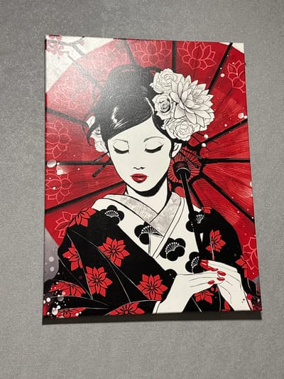A canvas print of a geisha is on a gray surface. The artwork depicts a geisha wearing a kimono with red flower patterns, lying under a red parasol with white flowers in her hair. Her hands are clasped, with red painted nails. The background of the parasol is red with a white floral pattern.