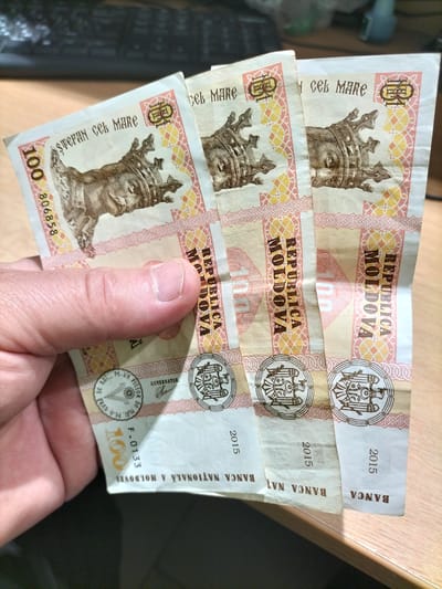 This close-up image shows a hand holding three Moldovan 100 Leu banknotes. The banknotes are predominantly cream and pale pink, with intricate designs featuring historical figures and national symbols. The portrait of Stephen the Great (Ștefan cel Mare) is prominently displayed on each note. The text "REPUBLICA MOLDOVA" and the denomination "100" are visible. The year "2015" is also printed on the lower section of each banknote. The background suggests an indoor setting, possibly a desk with a computer keyboard faintly visible. The lighting is artificial, casting a warm glow on the scene. The image focuses on the details of the currency, highlighting its design and texture. The hand holding the banknotes is positioned to showcase them, with a thumb and fingers gently grasping the edges. The overall impression is one of presenting or examining the currency. The text "BANCA NAȚIONALĂ A MOLDOVEI" confirms the origin of the banknotes. The location context of Telenești, Moldova, is provided, although not directly evident from the image itself.