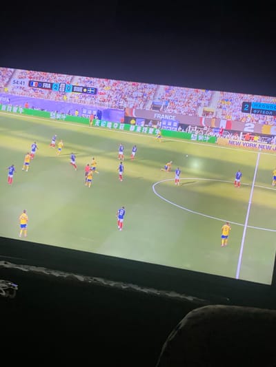 The image shows a soccer match displayed on a television screen. The score board indicates France is playing Sweden, with France leading 2-0. The time is 54:41. Players in blue and white uniforms (France) and yellow and blue uniforms (Sweden) are visible on the green field. The crowd in the stadium is blurred in the background. The television is positioned in a dark room, with a portion of the screen reflecting some ambient light. There is no indication of Zastavna, Ukraine in the image.