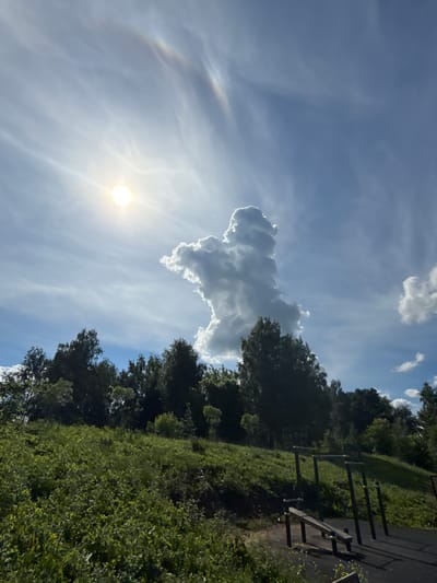 The image depicts a public outdoor fitness area on a grassy incline with a dense tree line at the top. The fitness equipment consists of parallel bars and a bench. The sky above is blue with scattered white clouds. A prominent cumulus cloud formation stands out in the center of the sky, resembling a figure. To the left, the sun is bright, with a subtle rainbow-like arc visible in the upper part of the sky, possibly indicating a circumzenithal arc or a sundog. The sun's rays cast shadows on the ground and equipment. The overall scene is one of a sunny day in a recreational park. The location is the city of Lysva, Russia.