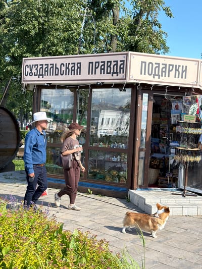 An outdoor scene in Suzdal, Russia, captures a bright, sunny day with a kiosk selling newspapers and gifts. Two people, a man in a blue denim shirt and white cowboy hat, and a woman in a brown t-shirt and baseball cap, walk past the kiosk. The man is holding a leash attached to a corgi dog walking in front of them. The kiosk has a sign reading "Суздальская Правда" (Suzdal Truth) and "Подарки" (Gifts). The kiosk displays various items, including magazines, trinkets, and other souvenirs. In the background, lush green trees and a clear blue sky create a pleasant atmosphere.