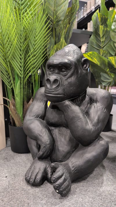 A black, resin statue of a seated gorilla is positioned in the foreground on a gray, carpeted floor. The gorilla statue is depicted in a pensive pose, with its chin resting on its left hand, and its right arm bent, resting on its leg. Its eyes are open, and its mouth is slightly downturned. Behind the statue, on the left side of the image, is a potted palm plant with vibrant green fronds. To the right of the palm, several large, artificial green leaves, some with yellow variegation, are visible. In the background, shelves stocked with items are dimly lit. The overall environment suggests an indoor retail space, potentially a garden center or home decor store, located in Sömmerda, Germany.